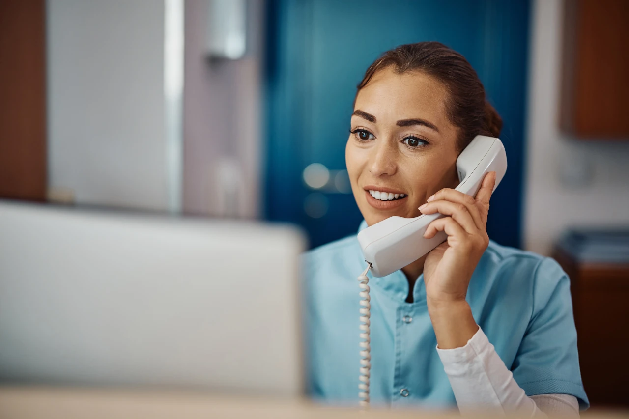 A lady speaking on a wired telephone while looking at a computer monitor and smiling