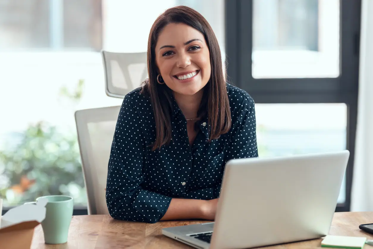 
                  A smiling woman sits at a desk with an open laptop in front of her
                