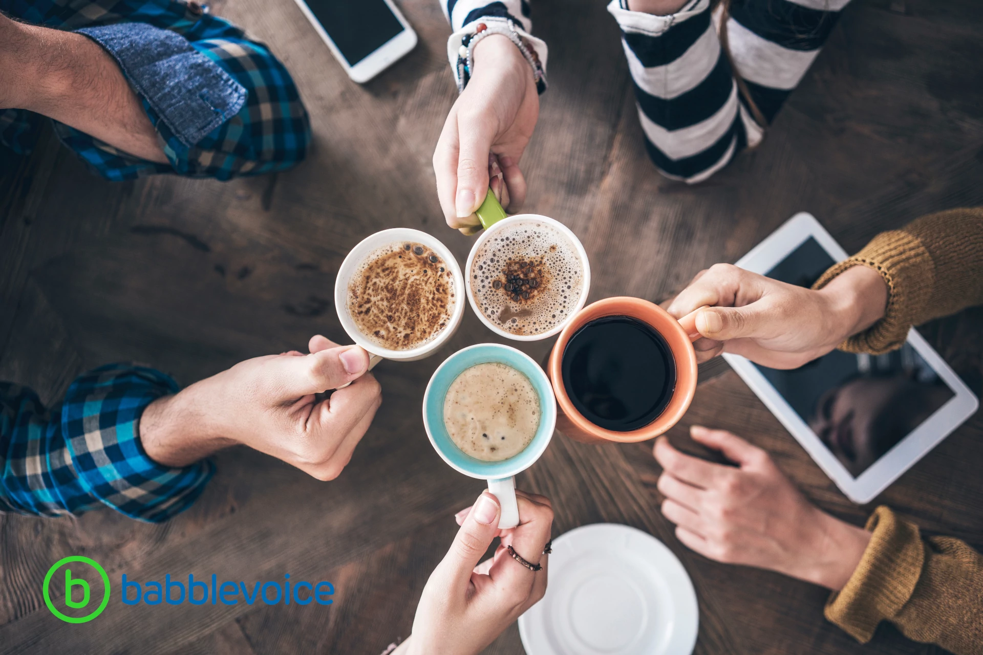 Four people hold coffee cups together over a table with phones and tablets on it.