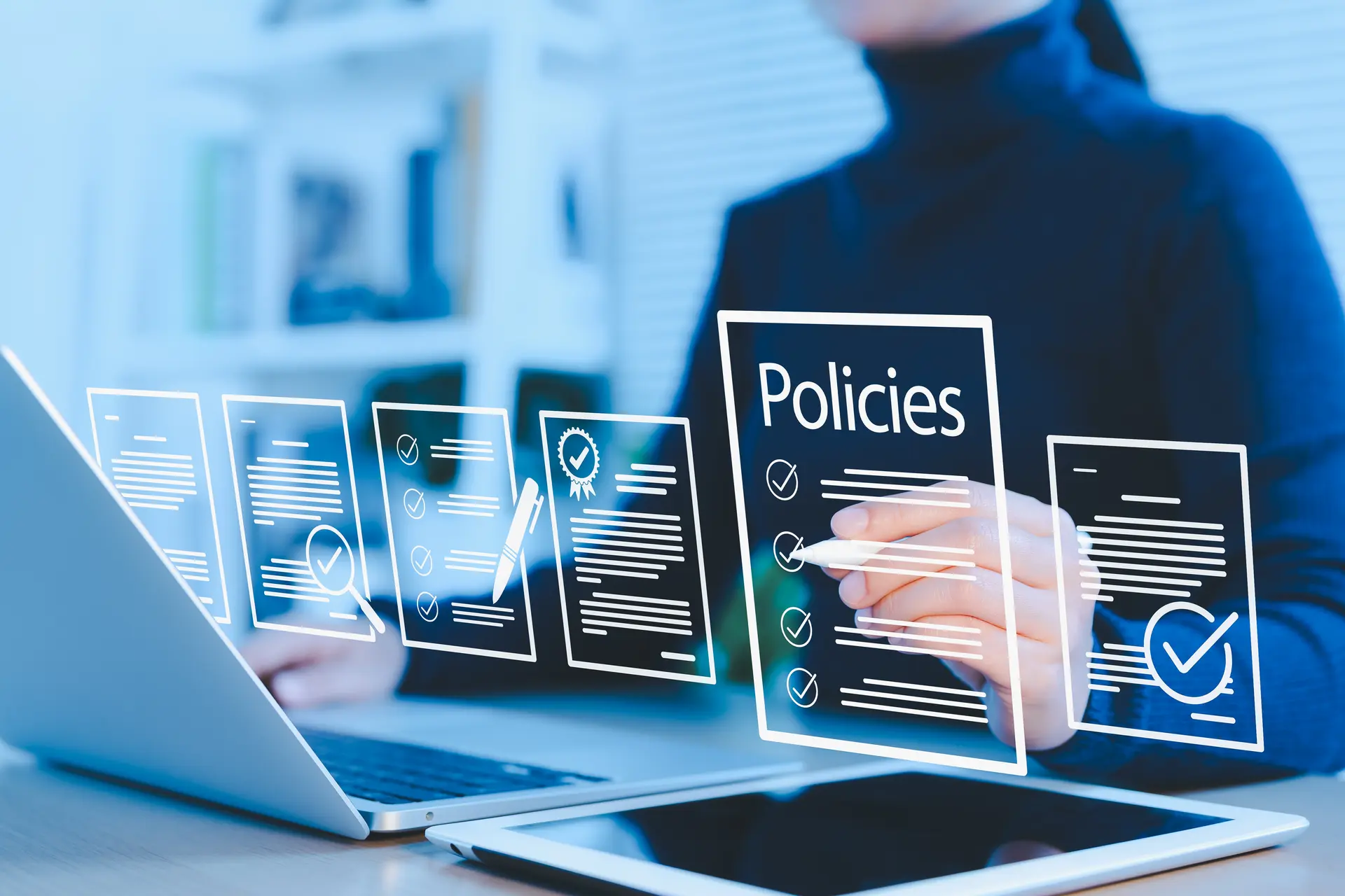 
                A person works at a desk with a laptop and tablet, reviewing a series of digital documents shown as a graphic overlay, including one headed 'Policies' with a row of ticked checkboxes.
              