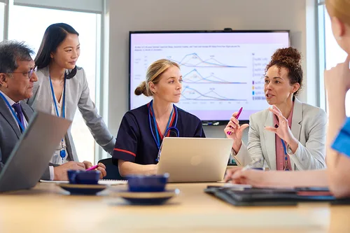 
                      A clinical team collaborating around a laptop during a meeting, with health data graphs displayed on screen behind them
                    